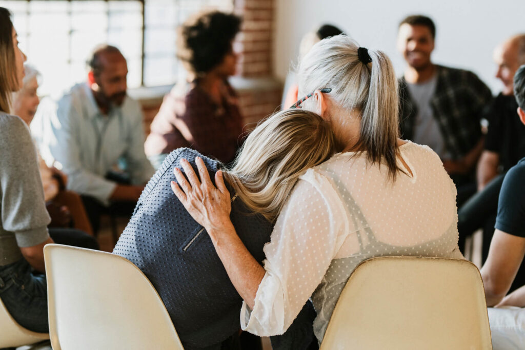 Two women comfort one another while sitting on chairs in a circle.